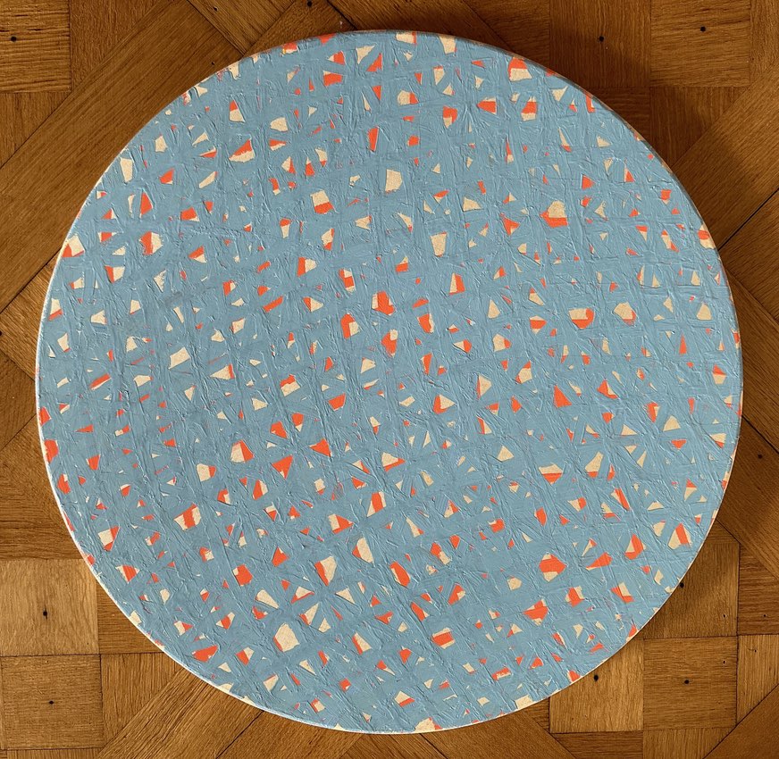 A circular canvas painting on a wooden floor, featuring a dynamic pattern of light blue background with scattered white and orange geometric shapes, suggesting a playful and modern artistic composition.